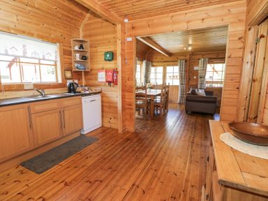 A wooden kitchen with cabinets a sink dishwasher and kettle next to a dining area with a table chairs and a sofa at Sun View Lodge Llanbedr