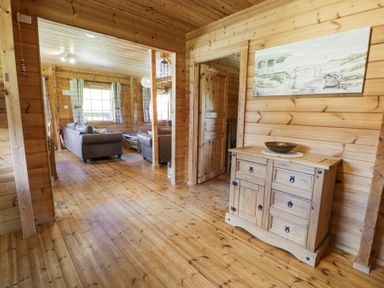 A wooden hallway with a wooden cabinet and a painting on the wall leading to a living room with sofas and a window at Sun View Lodge in Llanbedr