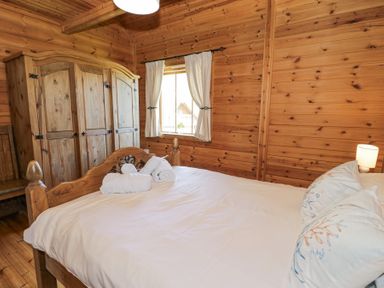 A bedroom with a wooden bed, wardrobe, chair, window with curtains, and bedside lamp at Sun View Lodge in Llanbedr