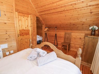 A bedroom with wooden walls ceiling and floor a bed with white linens and towels a wooden chair and a chest of drawers with flowers at Sun View Lodge in Llanbedr