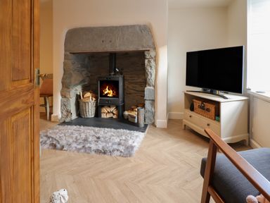 A living room with a wood burning stove in a stone fireplace with baskets of wood a TV on a stand and a chair at Bodlondeb in Tremadog