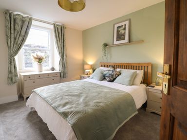 A bedroom with a double bed covered with green and white bedding beside two nightstands with lamps and a dresser under a window with green curtains at Bodlondeb in Tremadog