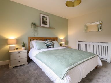 A bedroom with a bed with green comforter and pillows two nightstands with lamps and plants a decorative mirror on beige wall at Bodlondeb in Tremadog