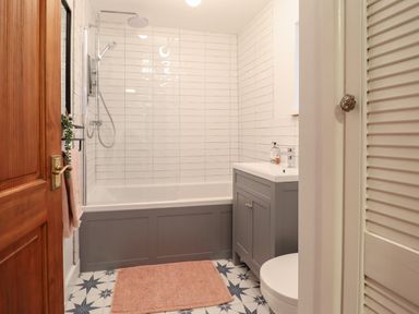 A bathroom with a bathtub shower combination a gray vanity unit a pink rug and patterned floor tiles at Bodlondeb in Tremadog