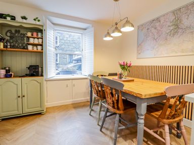 A dining area with a wooden table surrounded by chairs a cabinet with cups and a window with shutters at Bodlondeb in Tremadog