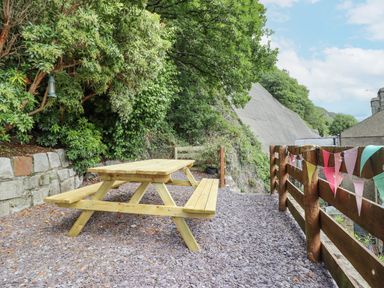 An outdoor seating area with a wooden picnic table surrounded by trees and a wooden fence with bunting at Bodlondeb in Tremadog