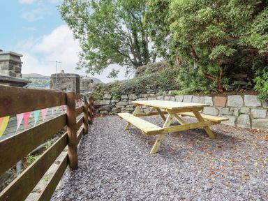 An outdoor area with a wooden picnic table on gravel next to a wooden fence and stone wall with trees at Bodlondeb in Tremadog
