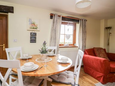 A dining area with a round wooden table set for four with white chairs next to a window and a red sofa at Lavender Cottage in Penruddock