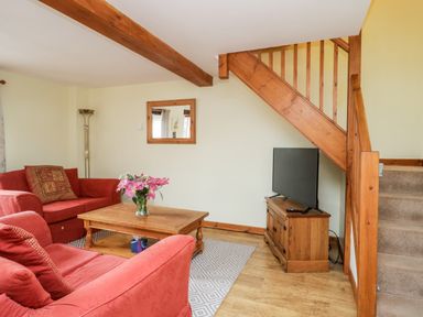 A living room with red sofas a wooden coffee table with flowers a television under wooden stairs at Lavender Cottage in Penruddock