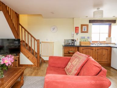 A living room with a red couch a wooden coffee table with pink flowers a television wooden stairs and a kitchen area at Lavender Cottage in Penruddock