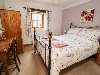 A bedroom with a bed with floral bedding a wooden wardrobe a desk with a chair and floral curtains at Lavender Cottage in Penruddock