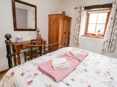 A bedroom with a floral bedspread pink and white towels on the bed a wooden wardrobe a window with floral curtains and a wooden desk with a chair and mirror at Lavender Cottage in Penruddock