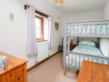 A bedroom with a bunk bed with floral bedding a wooden dresser a window with light curtains and a chair at Lavender Cottage in Penruddock