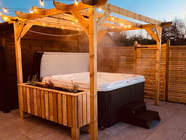 An outdoor hot tub under a wooden pergola with string lights a planter box and privacy wooden fencing at Daisy Cottage in Penruddock
