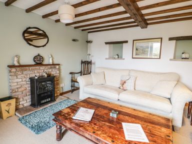 A living room with a beige sofa a wooden coffee table a brick fireplace and exposed wooden beams at Daisy Cottage in Penruddock