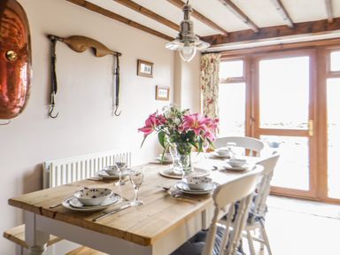 A dining room with a wooden table set with plates, bowls, glasses, and flowers with chairs and a glass door at Daisy Cottage in Penruddock