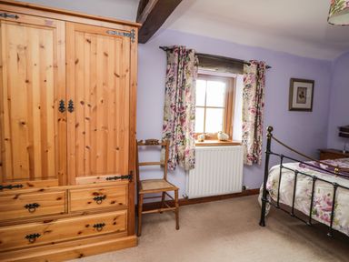 A bedroom with a wooden wardrobe a wooden chair floral curtains a metal bed frame and a window at Daisy Cottage in Penruddock