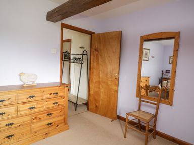 A bedroom with a wooden dresser a wooden chair wall mirror and an open door showing a clothes rack at Daisy Cottage in Penruddock