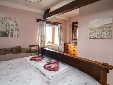 A bedroom with a wooden bed, folded towels on the bed, floral curtains, wooden dresser with mirror, and two paintings at Daisy Cottage in Penruddock
