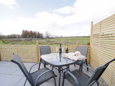 An outdoor patio with a glass table, four chairs, two bottles, glasses, and towels at Daisy Cottage in Penruddock