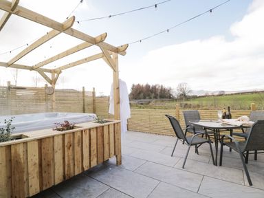 An outdoor patio with a hot tub under a wooden pergola and a table with four chairs overlooking a green field at Daisy Cottage in Penruddock