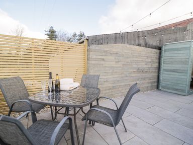 An outdoor patio with a glass table and four chairs and bottles with glasses on the table at Daisy Cottage in Penruddock