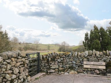 A stone wall with a wooden gate and a sign reading Daisy Cottage Parking with fields and trees in the background at Daisy Cottage in Penruddock