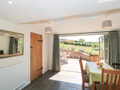 A dining area with a table covered in a green polka-dot tablecloth and wooden chairs next to open glass doors leading to a patio with outdoor furniture and garden at Lilac Cottage in Wookey Hole near Wells