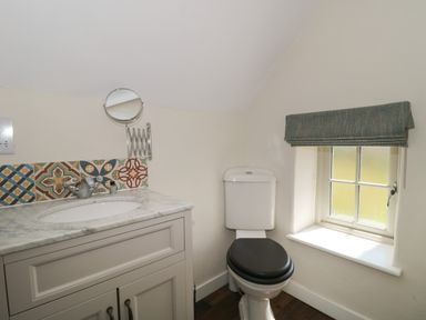 A bathroom with a toilet with a black seat cover a vanity with sink and patterned tile backsplash and a window with a fabric blind at Lilac Cottage in Wookey Hole near Wells
