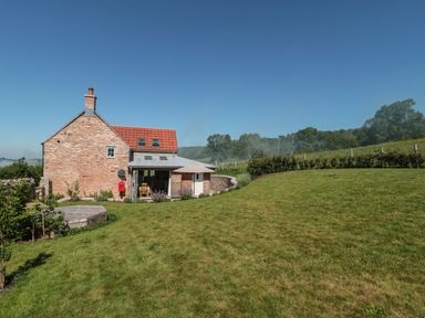 A stone house with a red roof and a large green lawn under a clear sky at Lilac Cottage in Wookey Hole near Wells