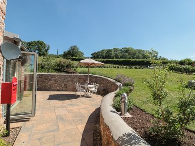 A patio with a table umbrella and chairs beside a brick wall and garden at Lilac Cottage in Wookey Hole near Wells