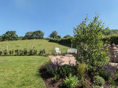 A garden with green plants and flowers next to a patio with white chairs and a bench at Lilac Cottage in Wookey Hole near Wells