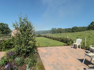 A garden with a lawn and chairs on a stone patio at Lilac Cottage in Wookey Hole near Wells
