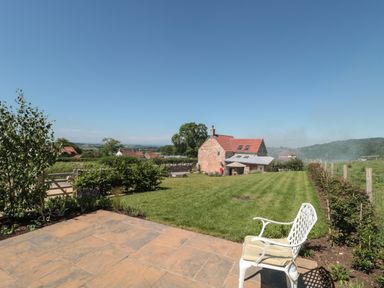 A garden with a white chair on a stone patio lawn shrubbery and a brick house in the background at Lilac Cottage in Wookey Hole near Wells