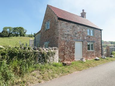 A stone house with a tiled roof next to a stone fence and a road at Lilac Cottage in Wookey Hole near Wells