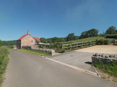 A rural road with a brick house with a red roof and stone walls next to a fenced garden and fields at Lilac Cottage in Wookey Hole near Wells