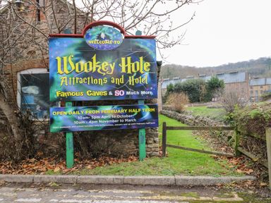 A sign for Wookey Hole Attractions and Hotel next to a grassy area with a wooden gate and stone buildings in the background
