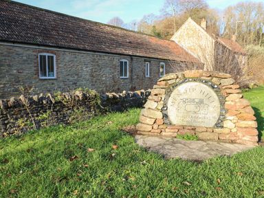 A stone sign for Mendip Hills Area of Outstanding Natural Beauty with stone buildings and grass around at Lilac Cottage in Wookey Hole near Wells