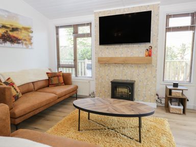 A living room with a brown sofa a wooden coffee table a yellow rug a wall mounted TV and windows at Broad Oak Lodge Applethwaite 7 Limefitt Park near Windermere