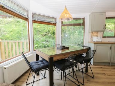 A dining area with a glass top table and six wireframe chairs next to large windows at Broad Oak Lodge in Applethwaite 7 Limefitt Park near Windermere