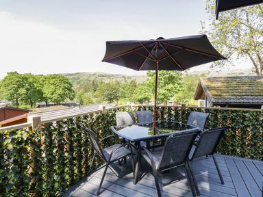 A balcony with a table and chairs under a large umbrella overlooking trees and rooftops at Broad Oak Lodge Applethwaite 7 Limefitt Park near Windermere