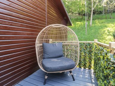 An outdoor balcony with a wicker chair with gray cushions next to a wooden wall and railing with green plants at Broad Oak Lodge Applethwaite 7 Limefitt Park near Windermere