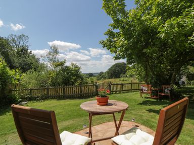 A garden with wooden chairs a round table with a flower pot on grass and a wooden fence at The Hideaway at Burrow Hill in Ottery St Mary