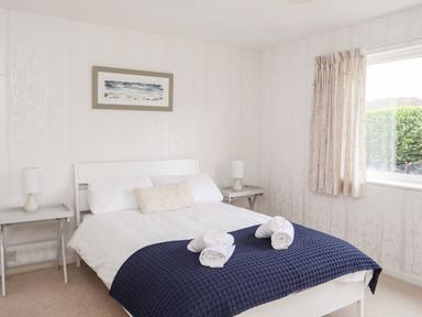 A bedroom with a double bed with white bedding and blue blanket two bedside tables with lamps and a window with curtains at Ty Bedw Benllech
