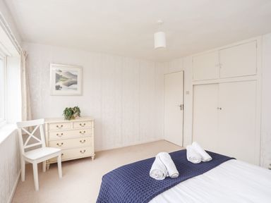 A bedroom with a bed covered in a blue blanket and white towels on it a white chair next to a cream chest of drawers with a green plant on top and a framed picture on the wall at Ty Bedw in Benllech