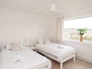 A bedroom with two single beds white linens pillows a chair between beds and a window with curtains and flowers at Ty Bedw in Benllech