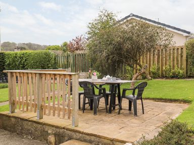 A backyard patio with a table and four chairs on a stone surface surrounded by grass and wooden fencing at Ty Bedw in Benllech