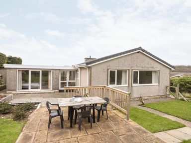 A backyard patio with a wooden table and plastic chairs outside a single-story house at Ty Bedw in Benllech