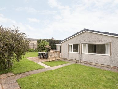 A backyard with green grass a paved area with a table and four chairs and a grey house with large windows at Ty Bedw in Benllech