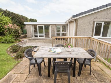 An outdoor patio area with a wooden table and four black plastic chairs next to a garden and a house with sliding glass doors at Ty Bedw in Benllech